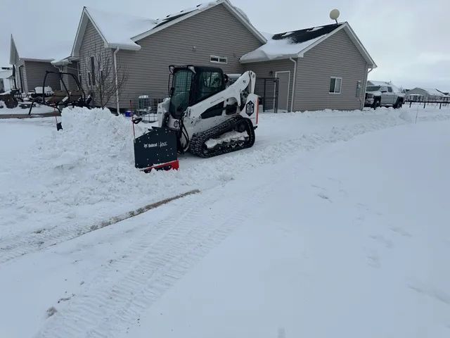 Snowy suburban driveway with a skid steer clearing snow beside a house
