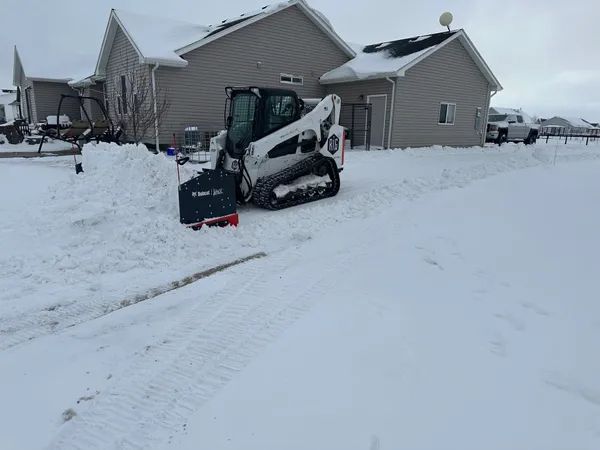 Snow-covered suburban driveway with a compact loader clearing snow beside a beige house.