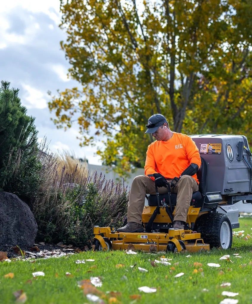 Person in bright orange shirt riding a yellow mower in a grassy yard with trees and flowers