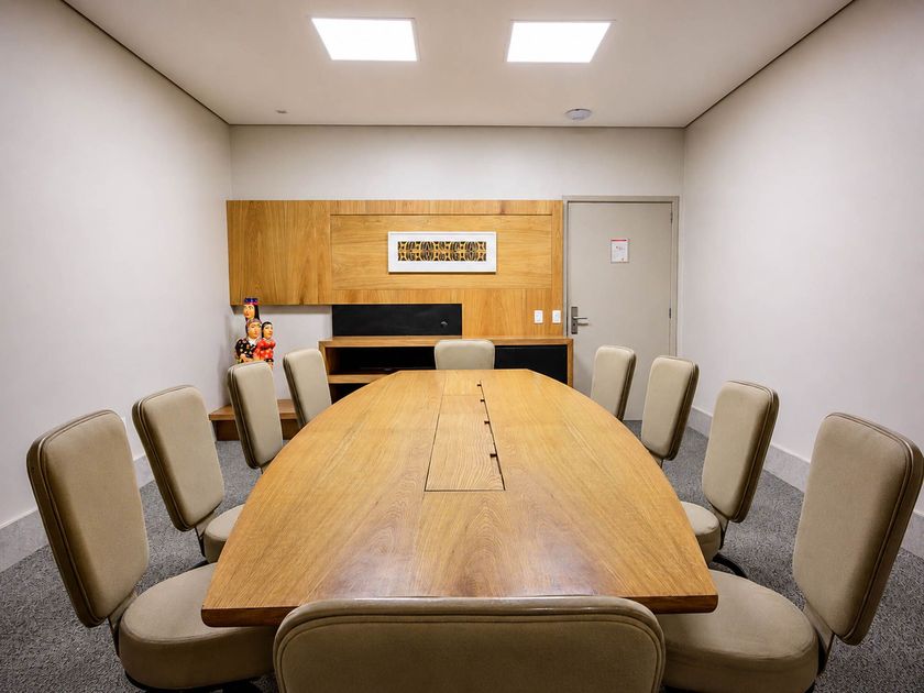 Sala de conferências vazia com uma longa mesa de madeira, cadeiras bege e um quadro na parede do fundo.