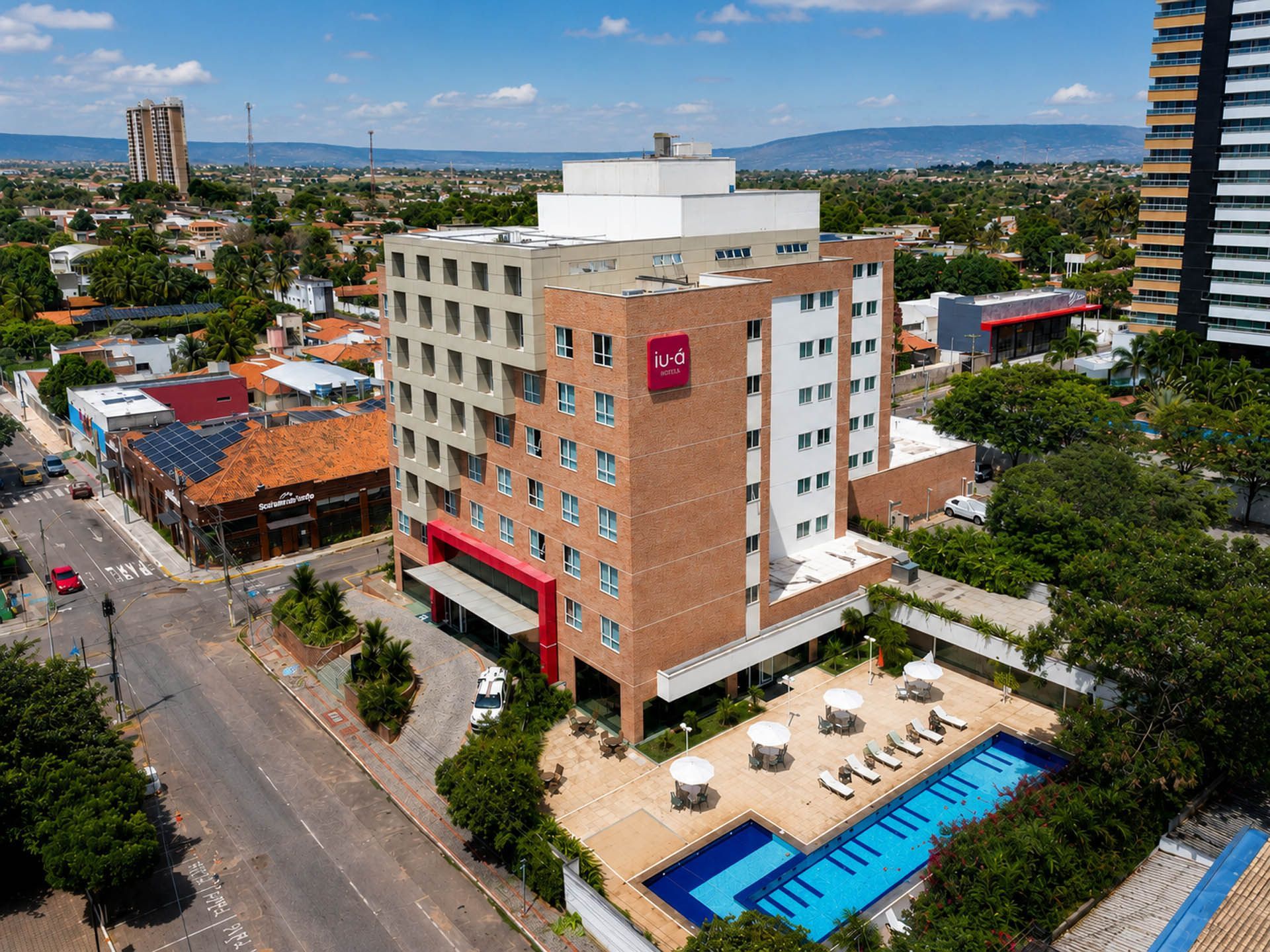 Vista aérea de um hotel de tijolos com uma piscina azul ao lado de uma rua da cidade e edifícios circundantes.
