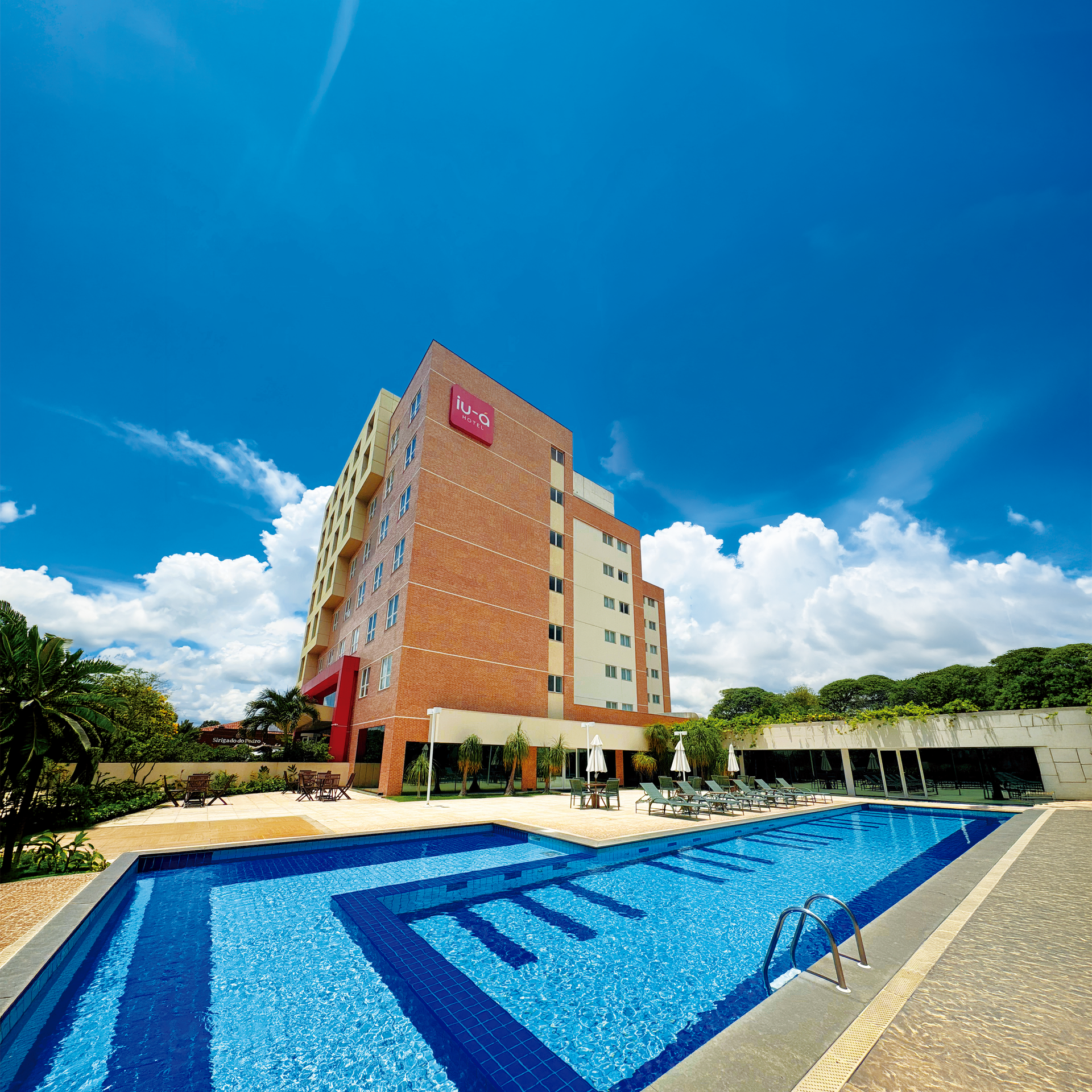 Vista aérea de um hotel de tijolos com uma piscina azul ao lado de uma rua da cidade e edifícios circundantes.