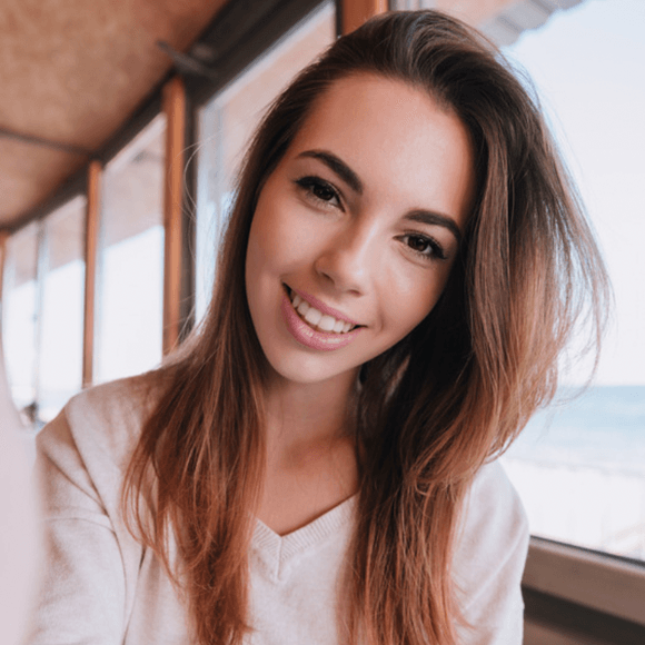 Woman with long brown hair smiles, inside by a window with a view of the ocean.