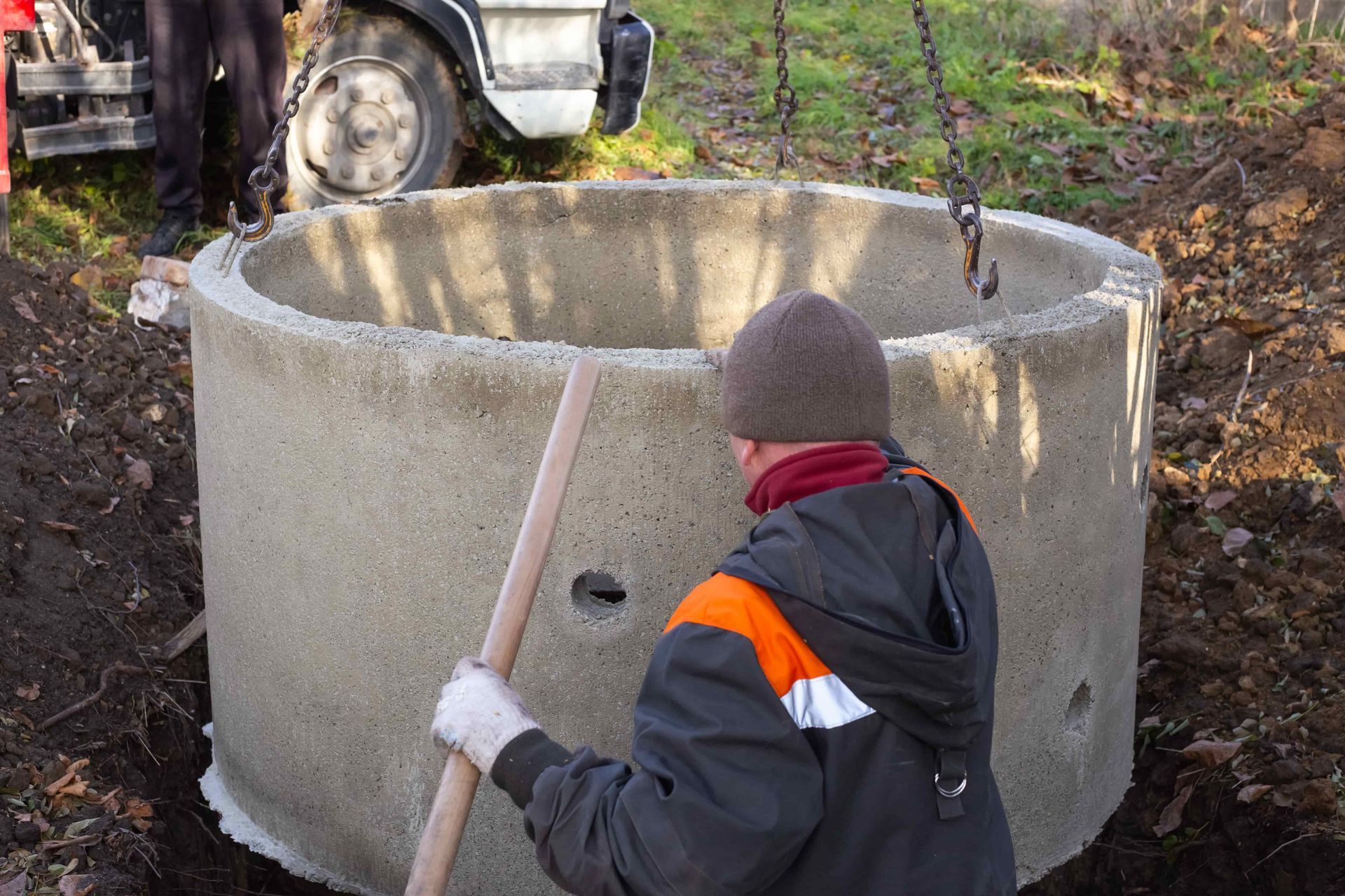 Man using a shovel near a concrete well ring being lowered by a chain, outdoors.