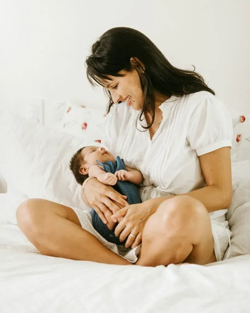 Woman sitting on a bed cradling a baby, looking down with a gentle smile.