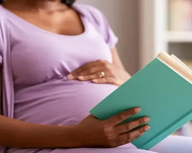 Pregnant person in a lavender shirt reading a teal book on a bed