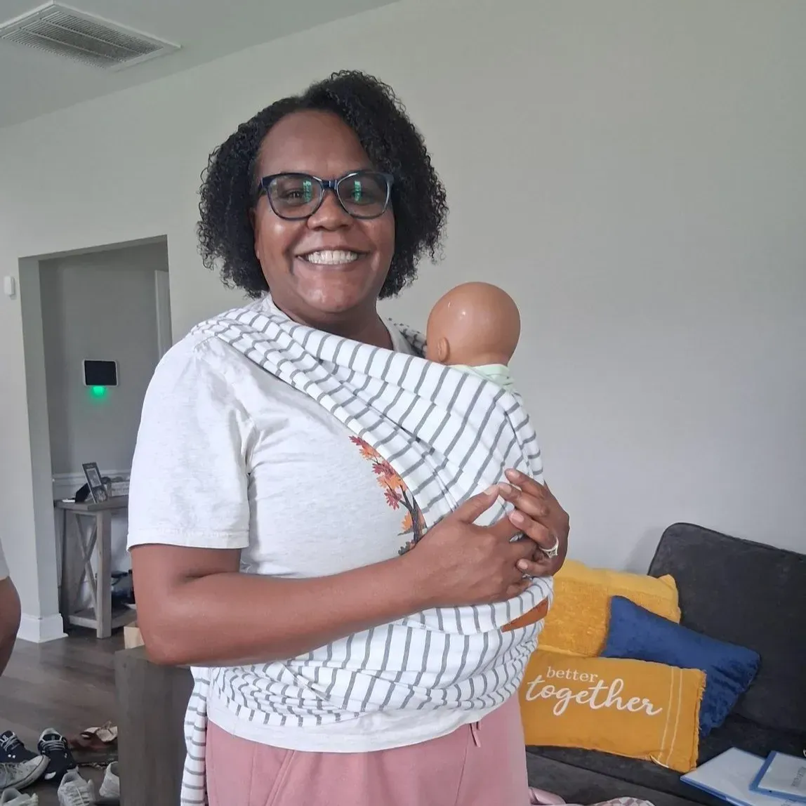 Person smiling while holding a baby doll in a living room, wearing glasses and a white shirt.