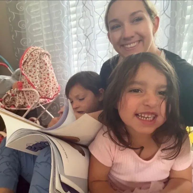 Smiling adult with two children reading a book on a couch by a window