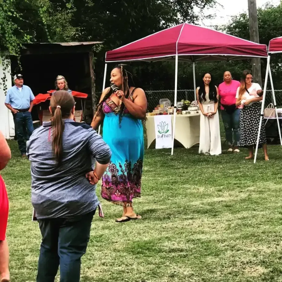 Outdoor community gathering with people standing under pink canopies on a grassy lawn, one woman speaking with a microphone.