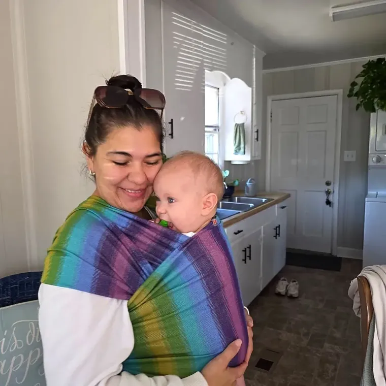 Woman smiling while holding a baby in a colorful wrap in a bright kitchen
