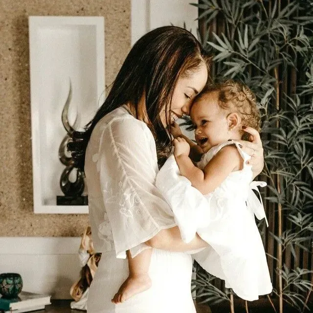 Woman holding a smiling child indoors, both in white clothing, sharing an affectionate moment