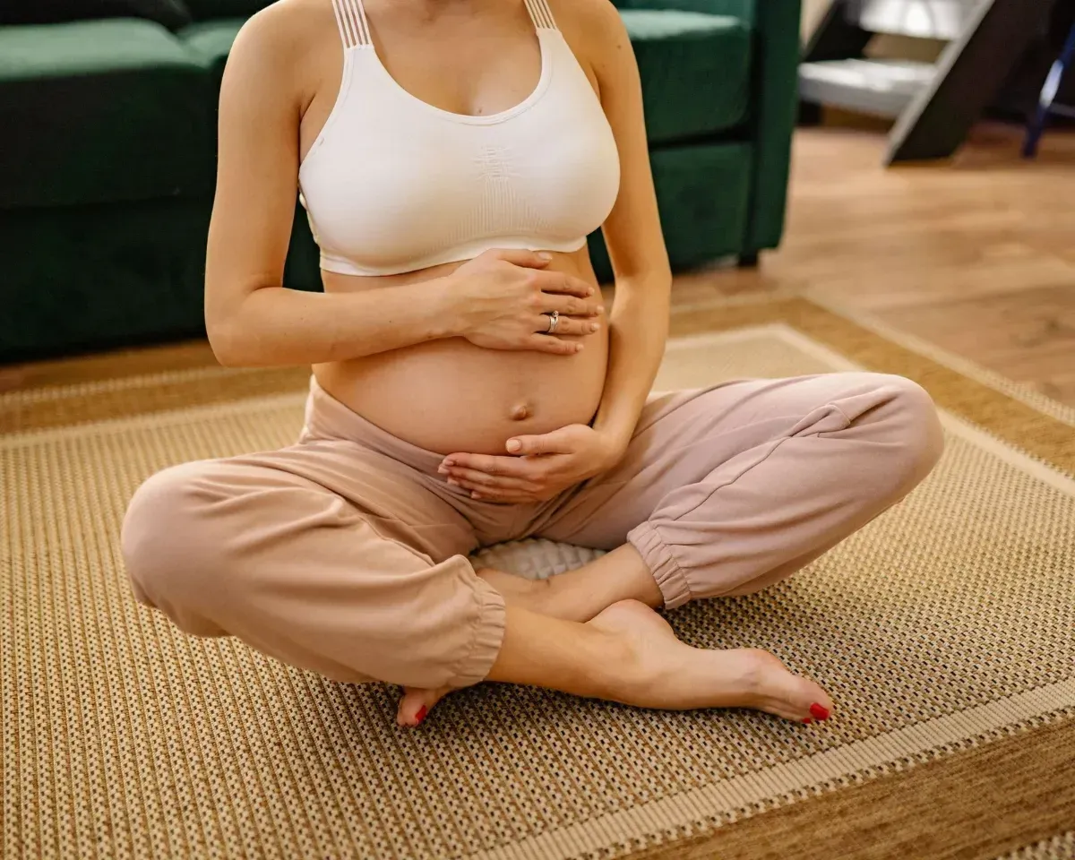 Pregnant person sitting cross-legged on a rug, holding their belly in a living room