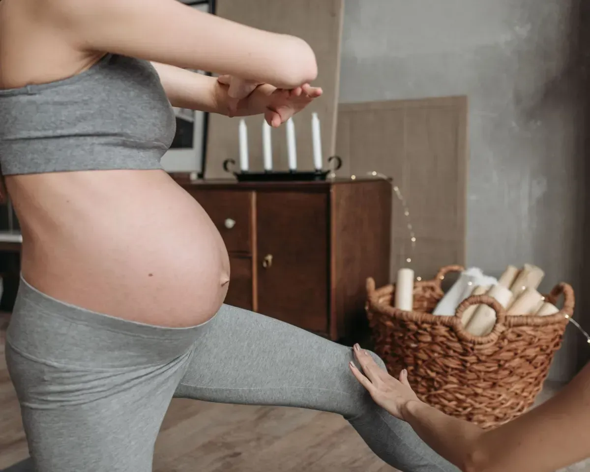 Pregnant person stretching a gray legging over a large belly in a bedroom