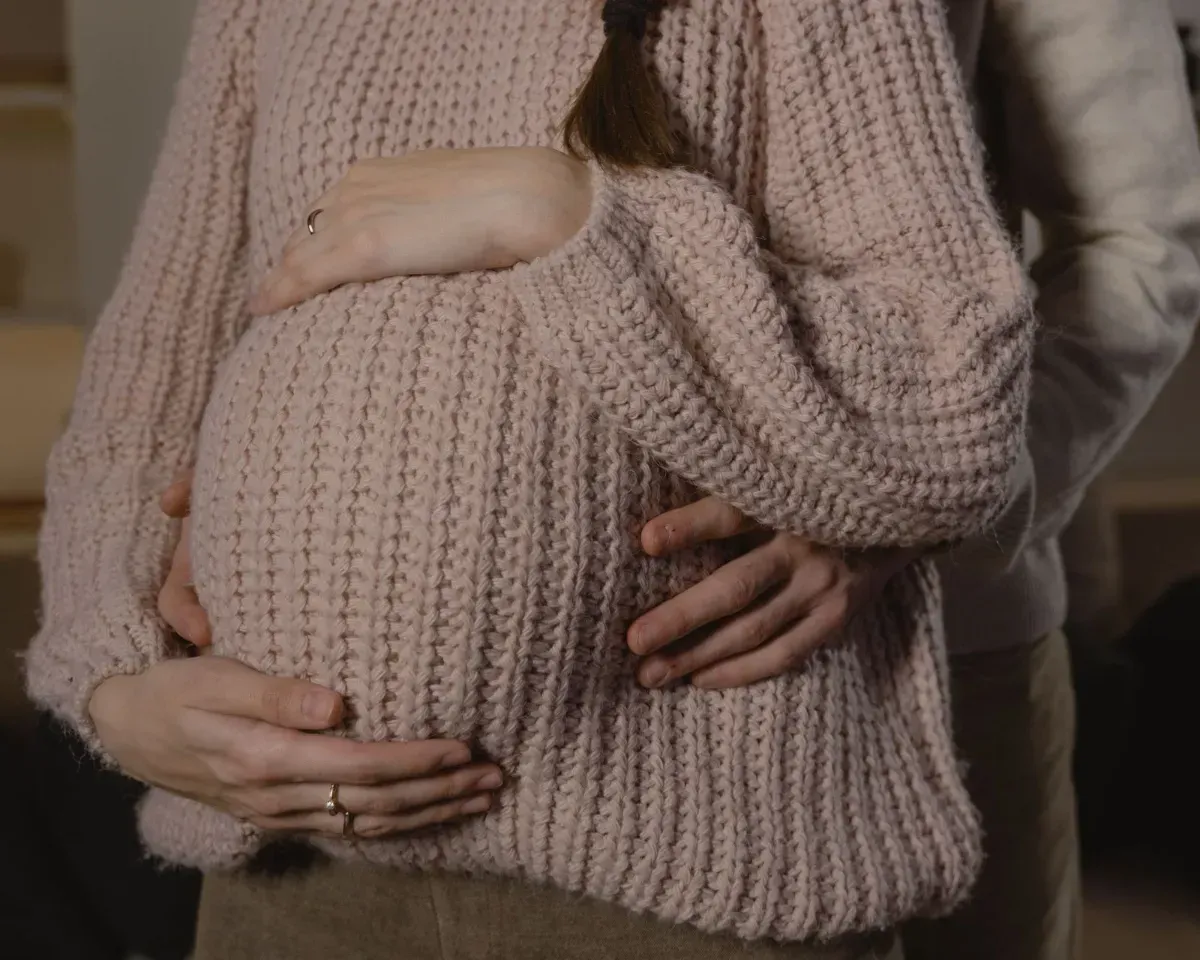 Hands on a pregnant belly under a chunky pink sweater in a softly lit indoor setting