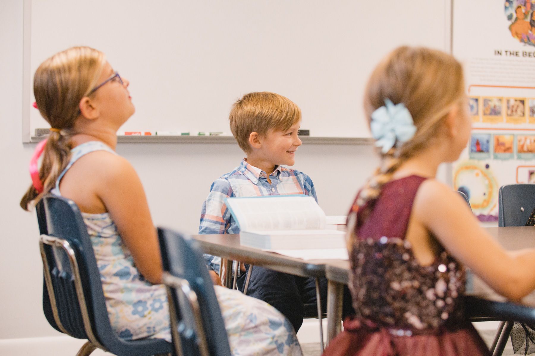 Three children seated at a table in a classroom; one girl looks up, the other looks forward, and a boy smiles.