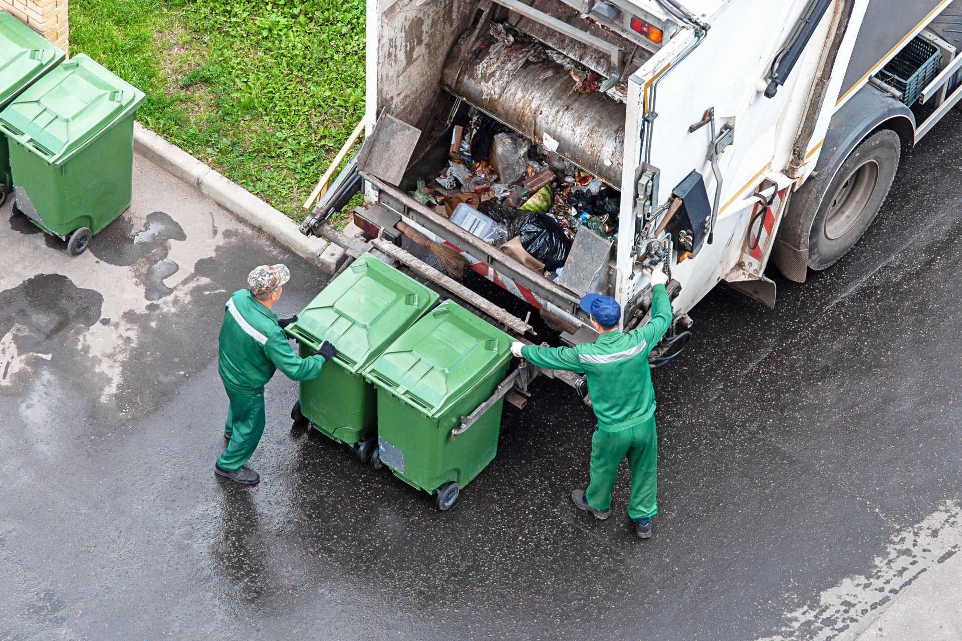 Two sanitation workers emptying green trash bins into a garbage truck on a wet street.