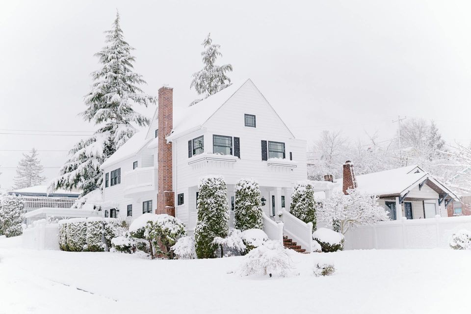 White house covered in snow with black shutters and brick chimney. Snowy trees and a smaller house in the background.