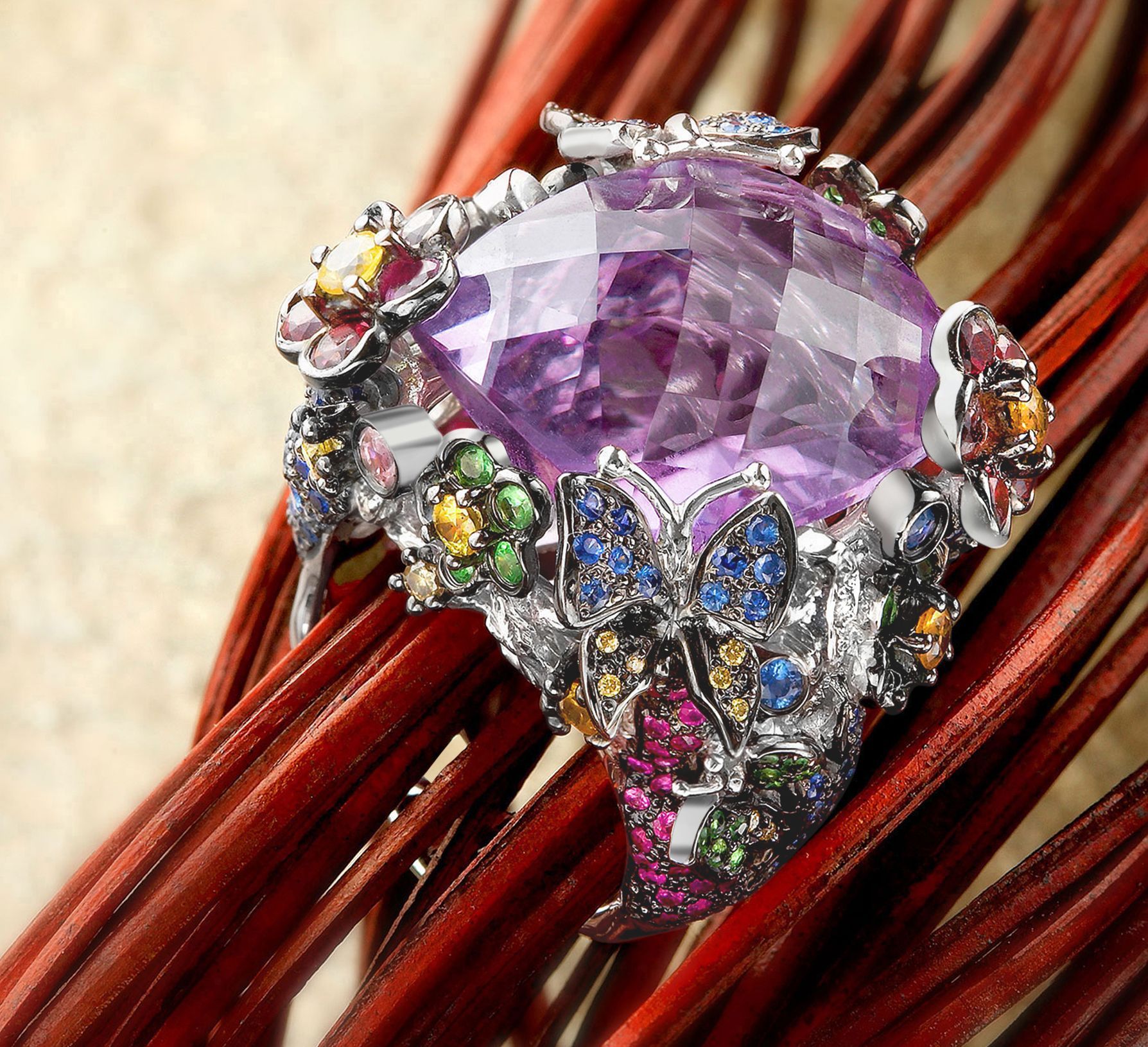 A large amethyst gemstone ring surrounded by colorful gemstones and floral accents, resting on red sticks.