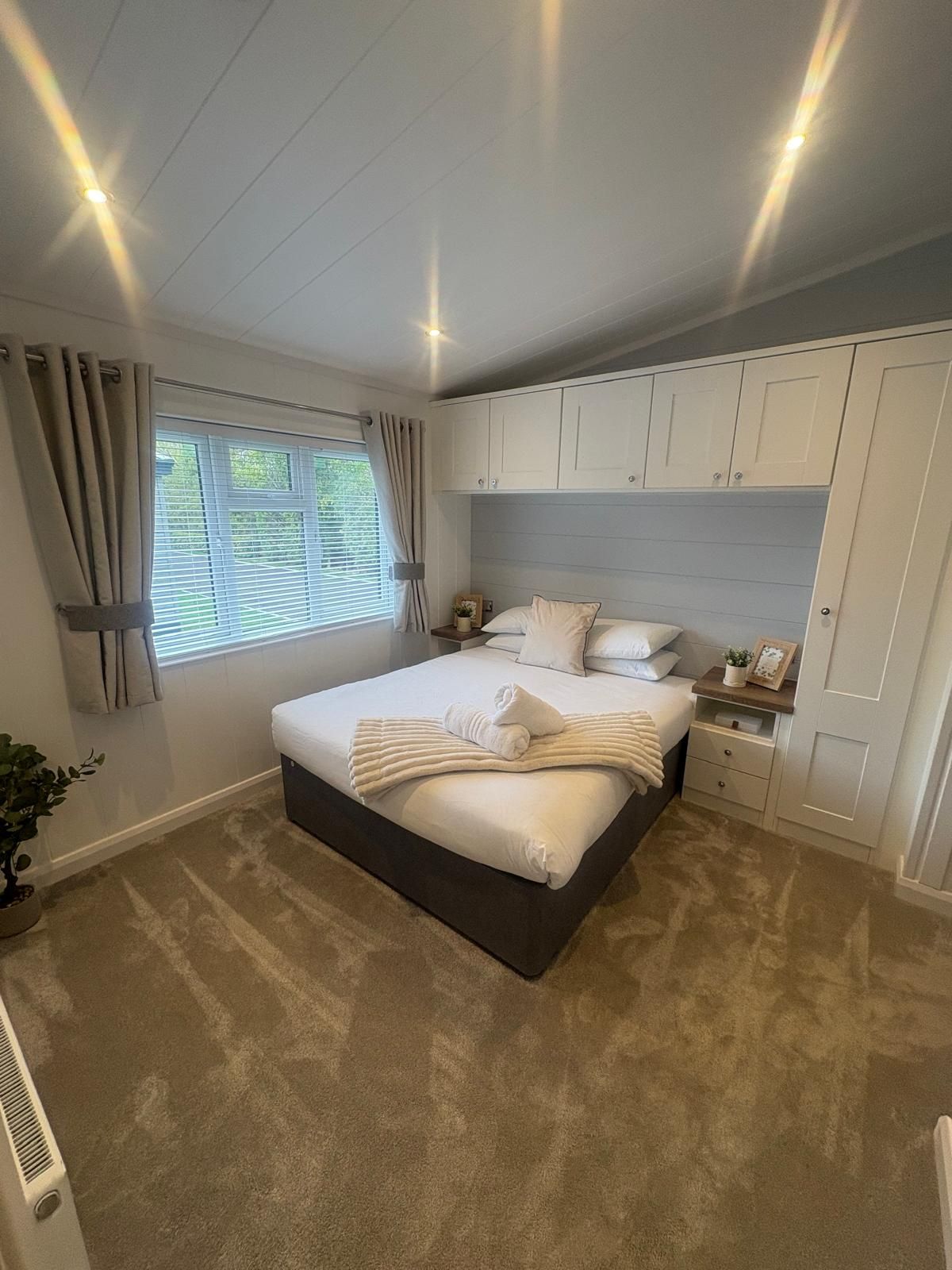 A neutral-toned bedroom with a double bed, overhead white storage cabinets, a window with curtains, and carpet flooring.