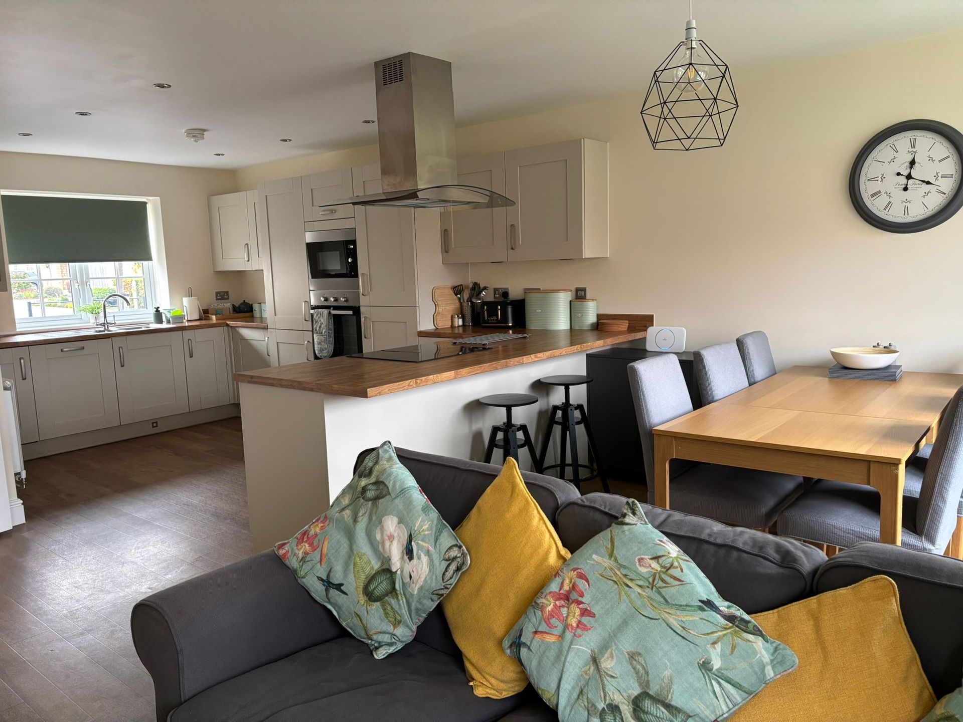 An open-plan kitchen and dining area with a grey sofa, wooden table, and light-colored cabinets.