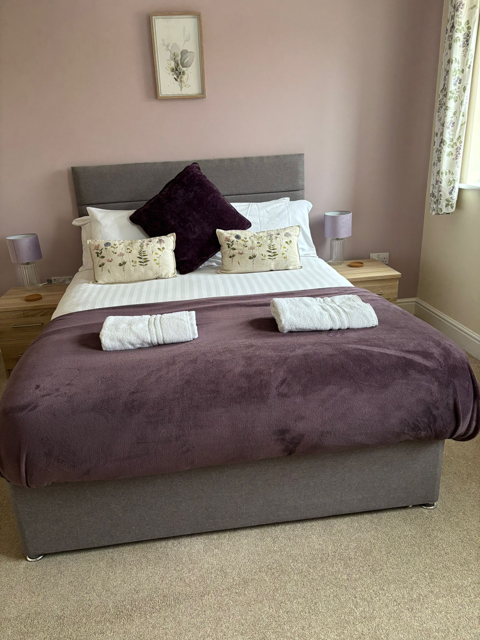 A tidy bedroom featuring a grey upholstered bed with a purple throw, floral accent pillows, and matching bedside lamps.