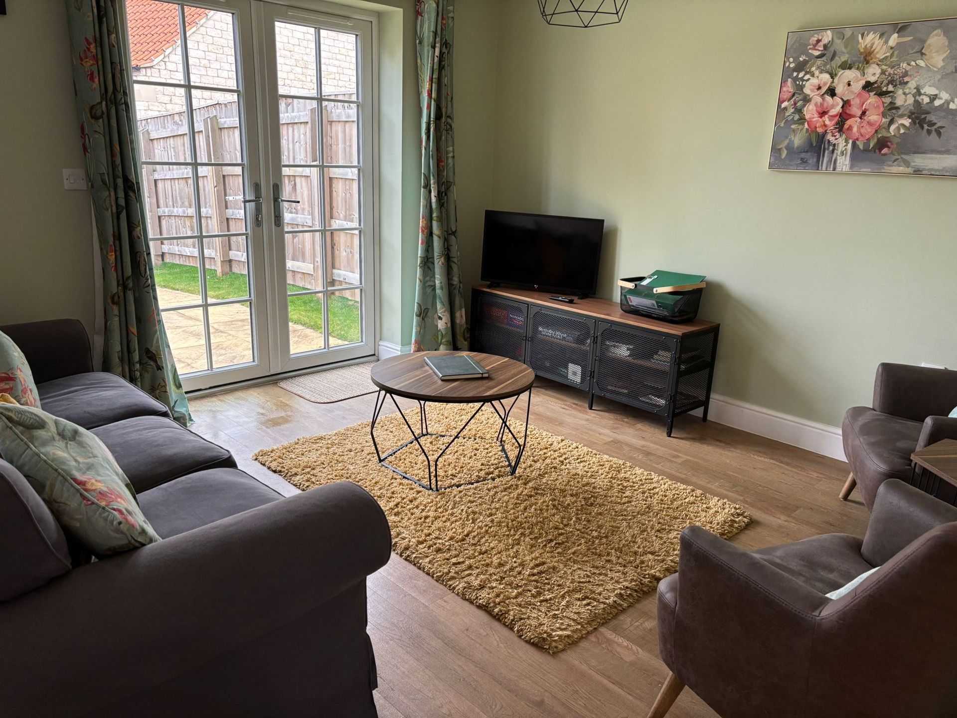 A cozy living room with a grey sofa, two armchairs, a round coffee table on a yellow rug, and a TV on a rustic unit.