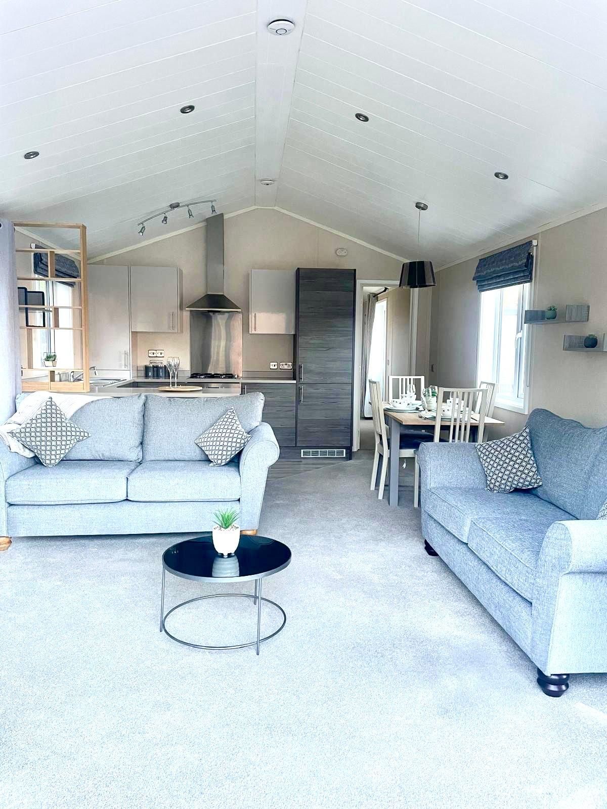 A bright, modern open-plan living room with two gray sofas, a round coffee table, and a kitchen area in the background.
