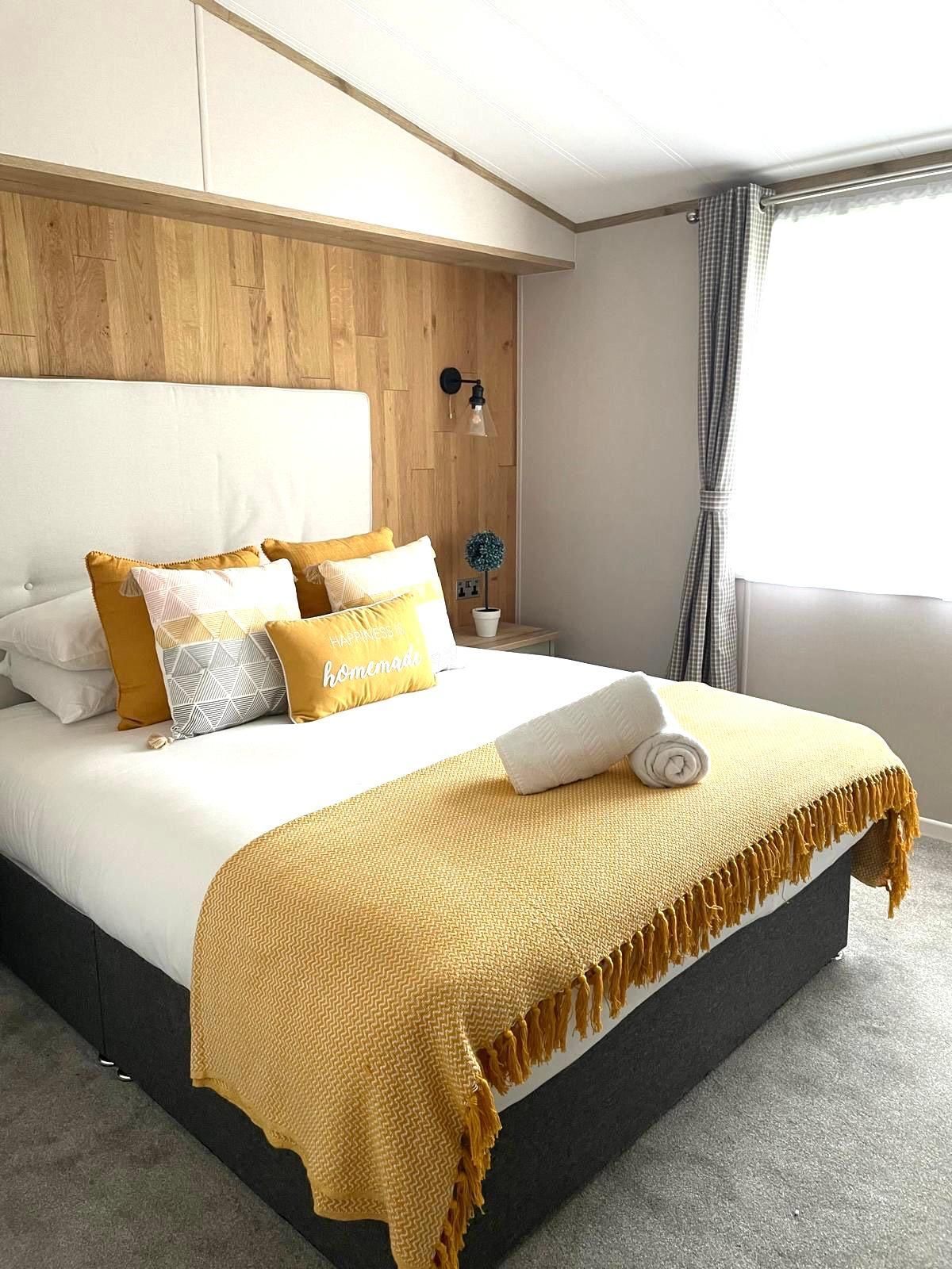 A bedroom with a queen-sized bed, white linens, yellow accent pillows, a yellow knitted throw, and wood paneled headboard.
