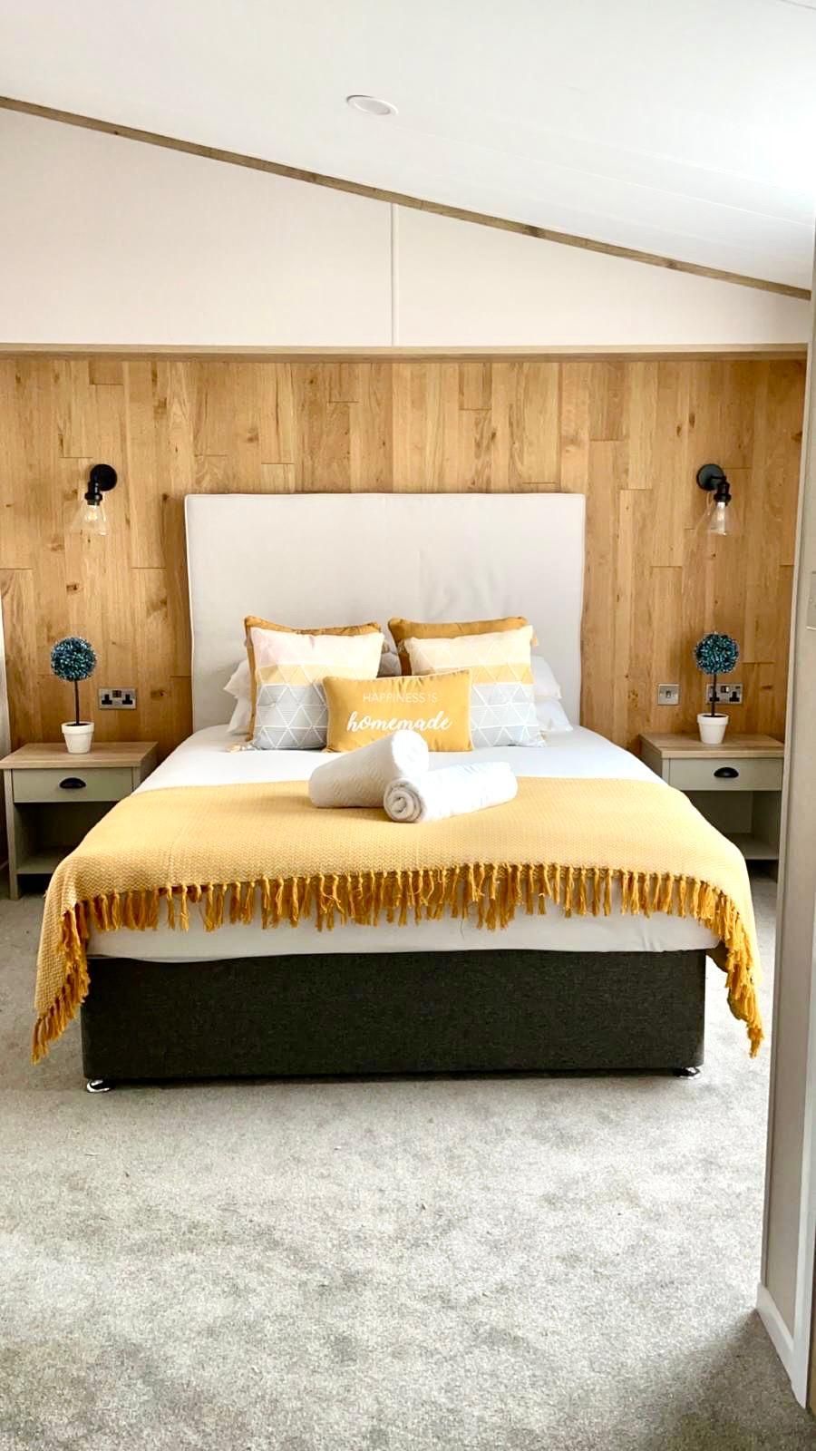 A bedroom featuring a bed with a white headboard, yellow throw blanket, pillows, and wood-paneled walls with bedside lamps.
