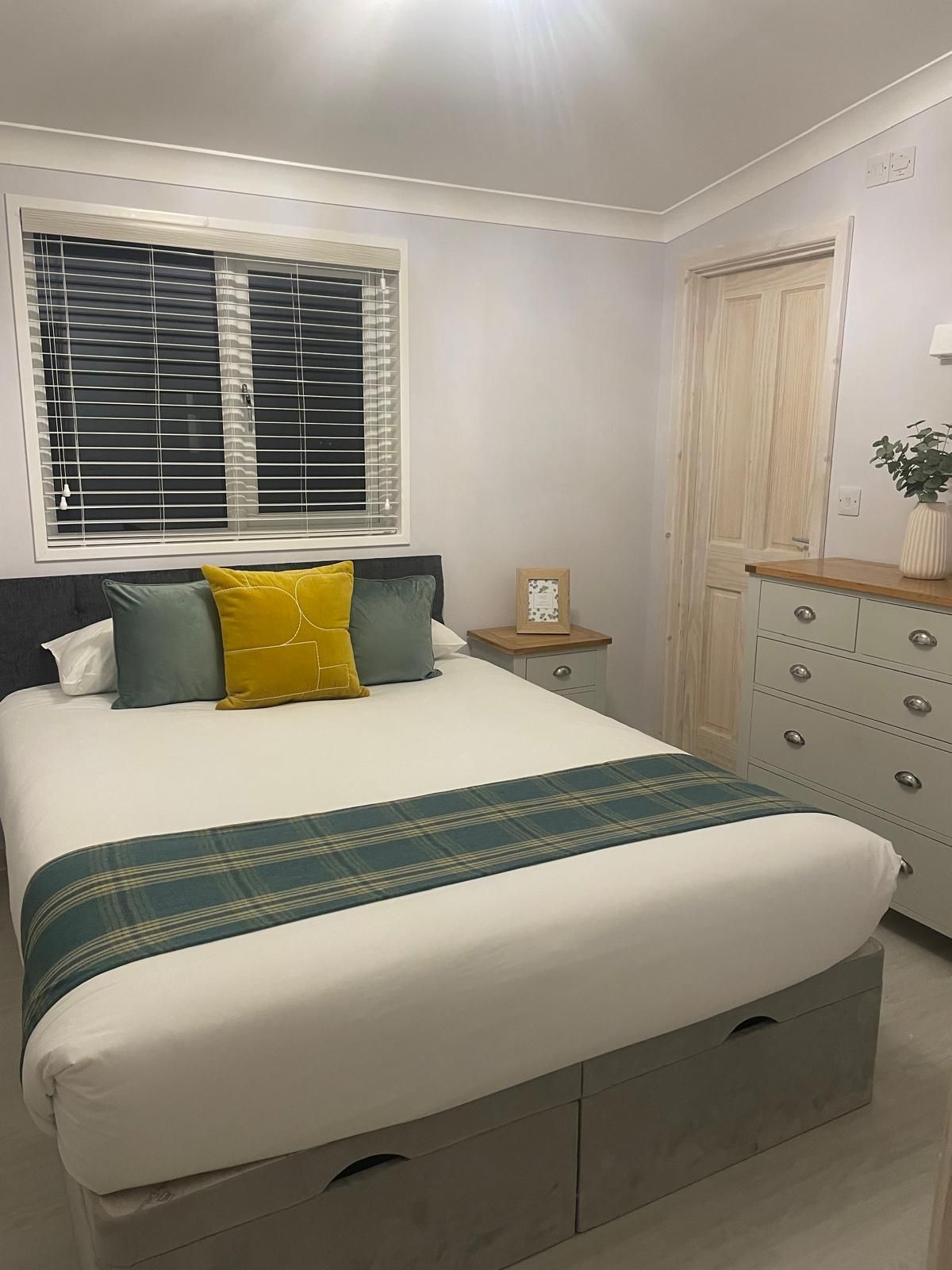 A bedroom with a queen bed featuring a white duvet, teal and yellow pillows, a matching runner, and a light wood dresser.