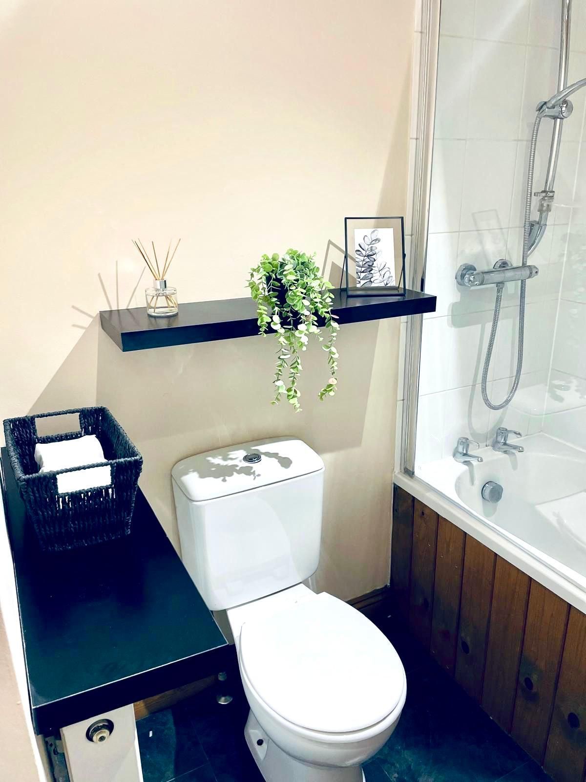 A bathroom features a toilet next to a black shelf with a small plant and a picture frame, near a tub with a wood panel.