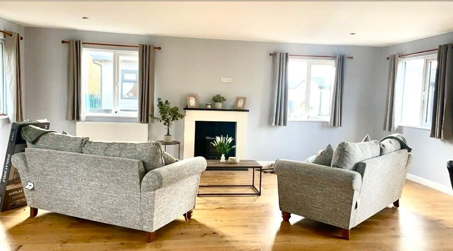 A spacious, sunlit living room with light gray walls, two gray sofas, a fireplace, and light wood flooring.