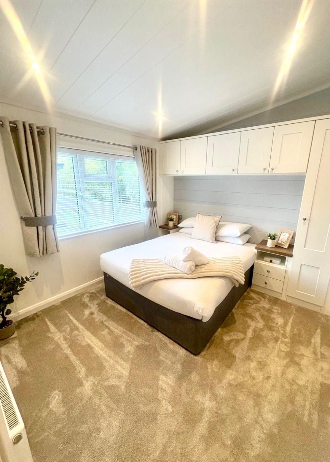 A cozy bedroom with a neatly made bed, light-colored walls, patterned carpet, and built-in white cabinets above the bed.