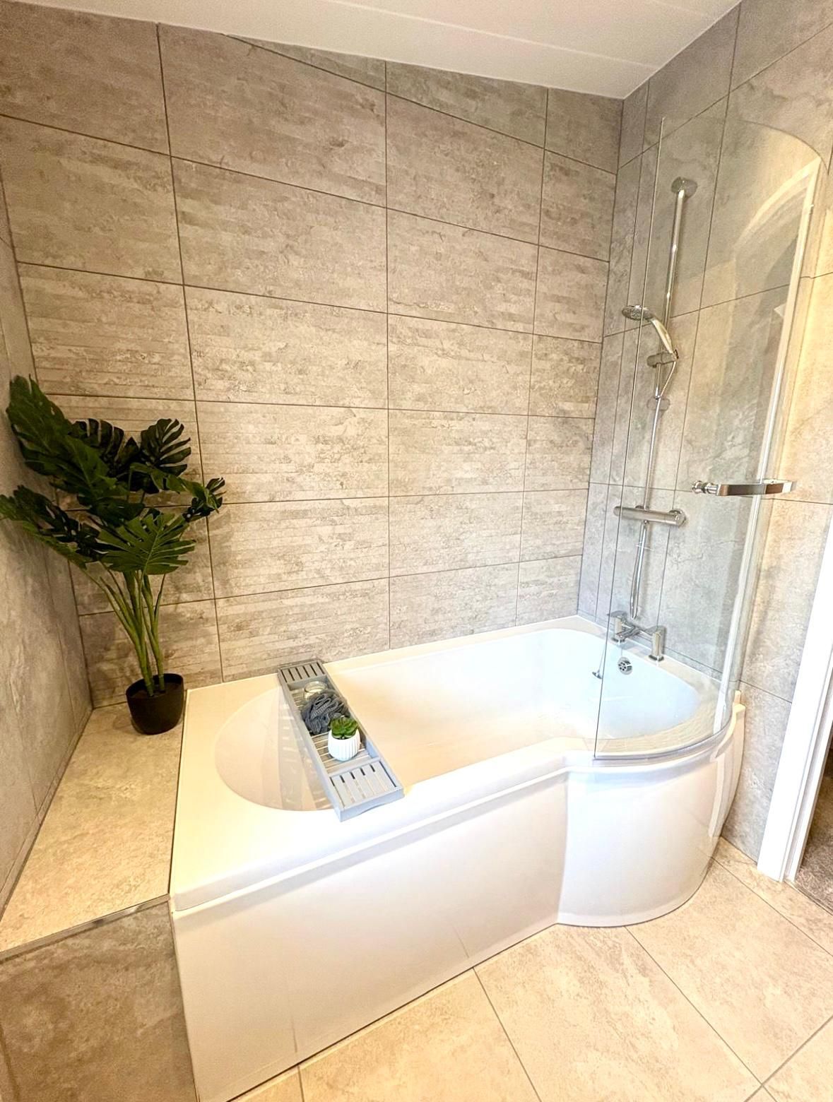 A white L-shaped shower-bathtub with grey stone-effect tiled walls and a potted plant on a built-in ledge.