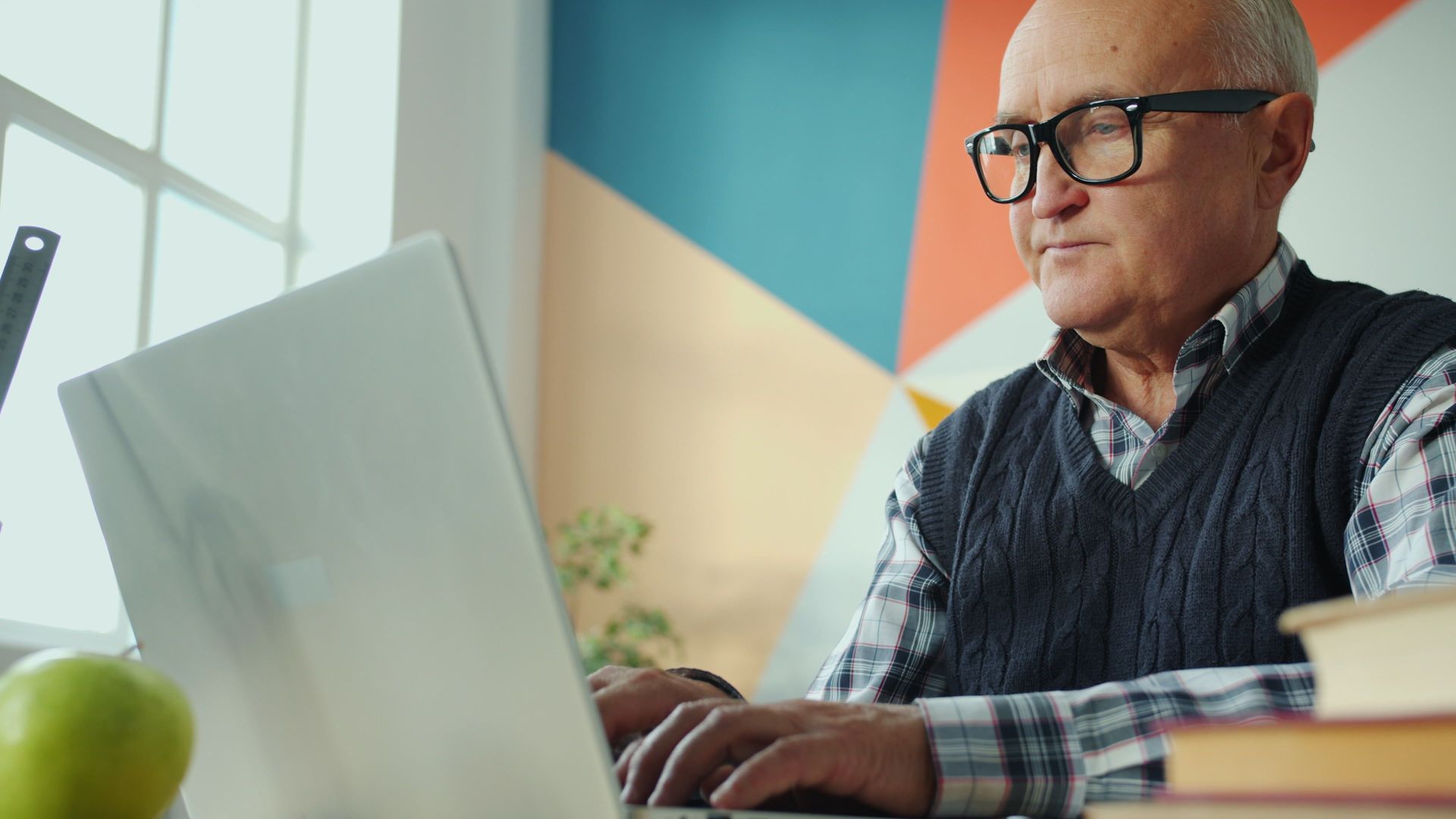 Man typing on a laptop at a desk, with a geometric wall backdrop and a green apple nearby