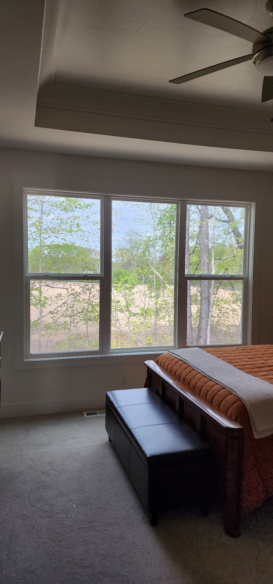 Bright bedroom with large windows, ceiling fan, gray carpet, and a bed with orange bedding.
