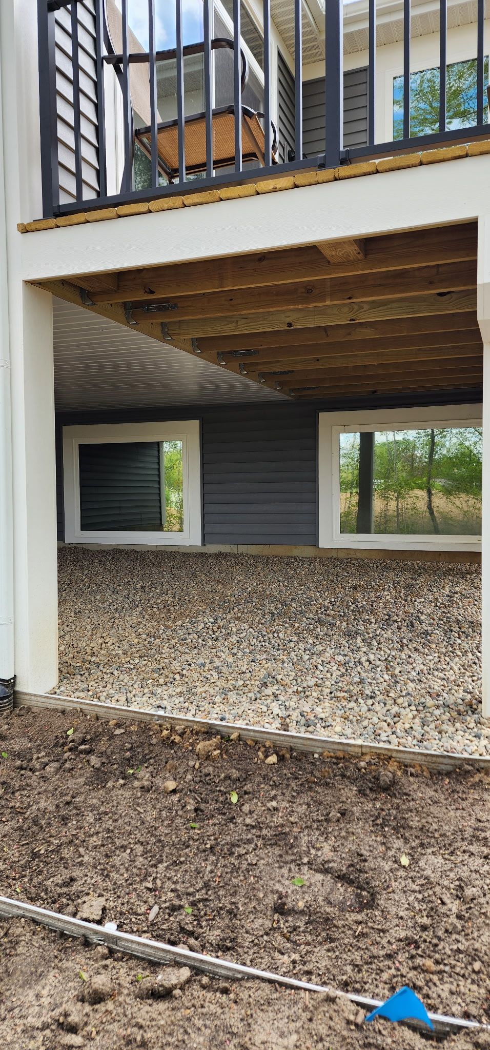 Ground-level view of a house exterior with a raised wooden deck, gray siding, and gravel-covered yard.