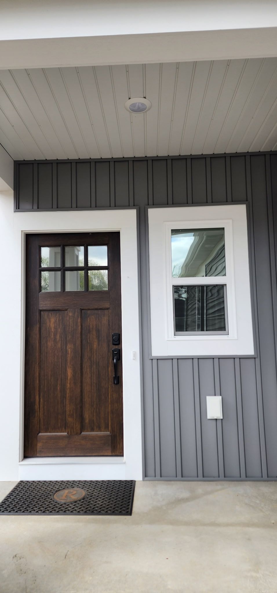 Front porch with a dark wooden door, white-framed window, gray siding, and a doormat.