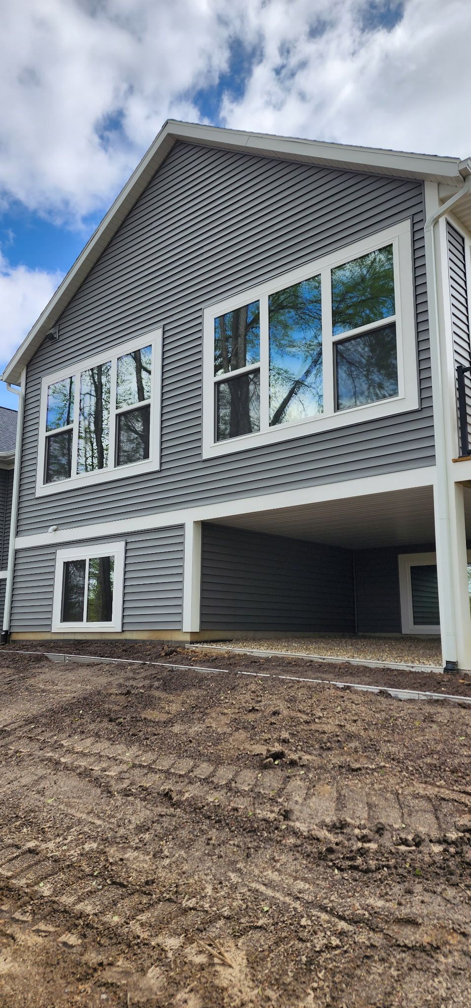 Newly built gray house with white trim and large windows on a dirt lot under a cloudy sky
