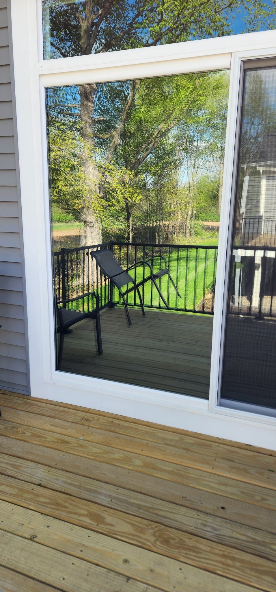 White-framed glass patio door reflecting a green yard, beside a wooden deck and gray siding.