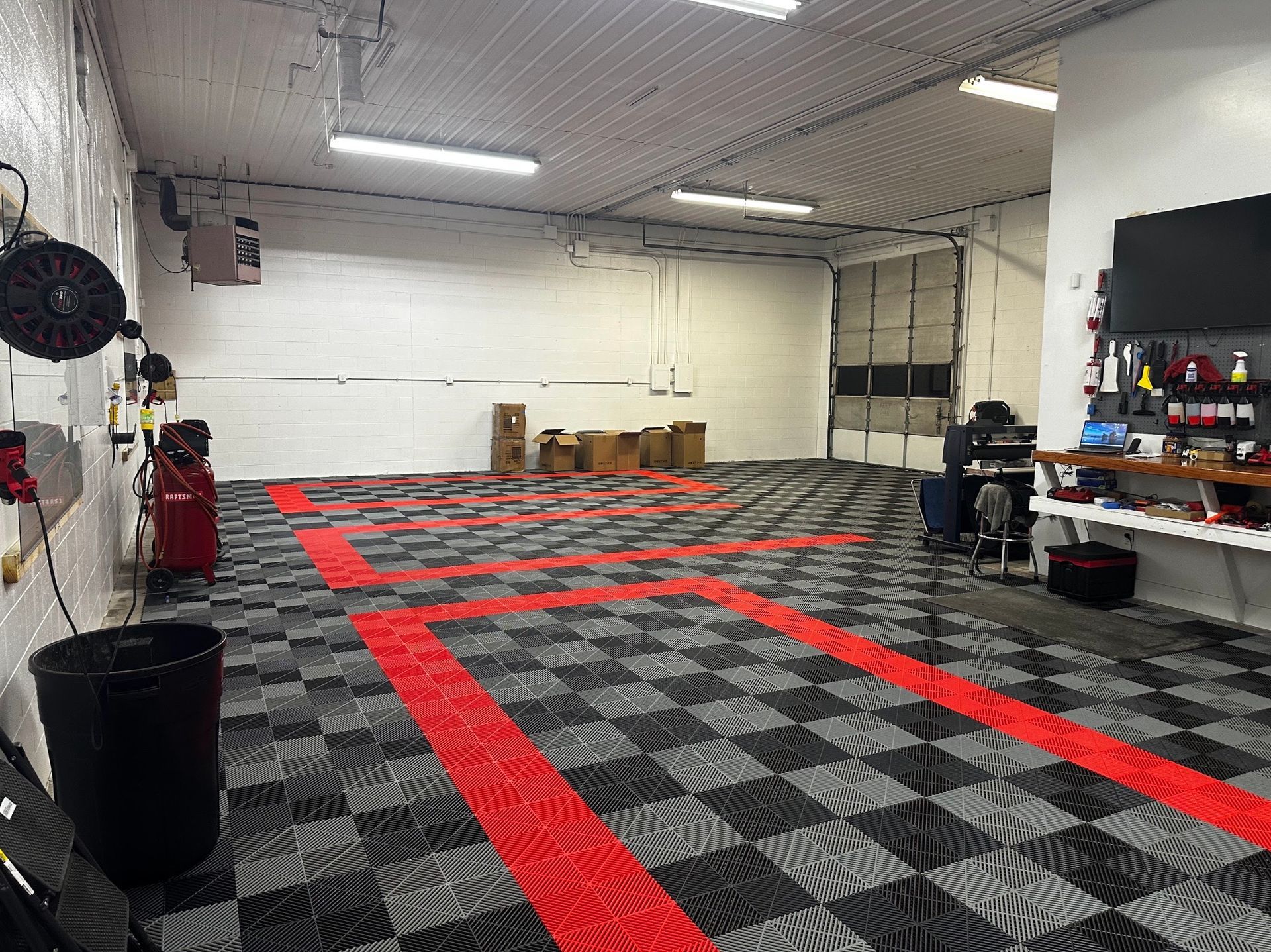 Spacious garage with gray patterned floor, red taped lanes, workbenches, and stacked boxes under fluorescent lights