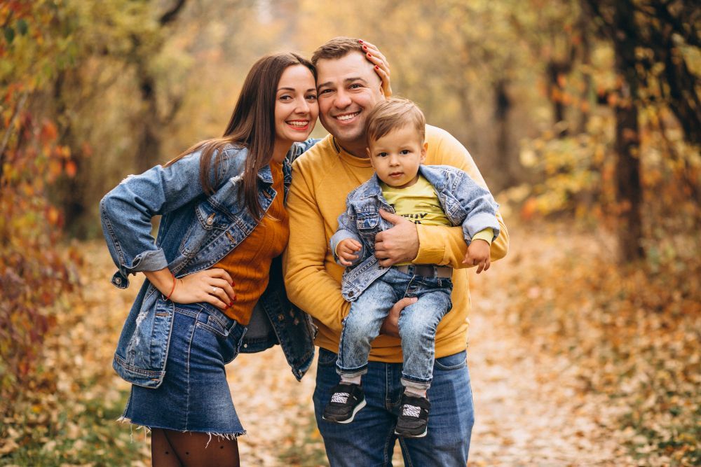 Three people smiling outdoors in autumn, with two adults and a child in denim and mustard clothing.