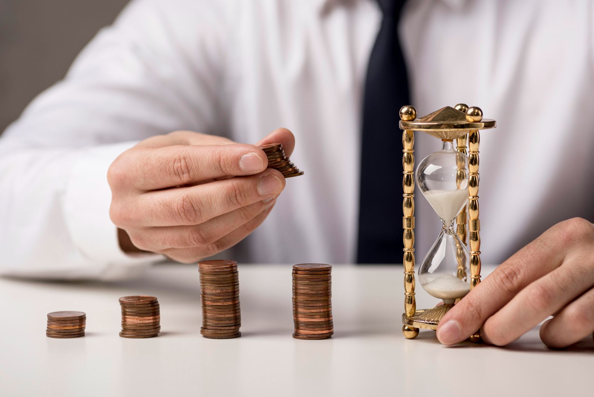 Person stacking coins beside an hourglass on a white table, symbolizing time and savings