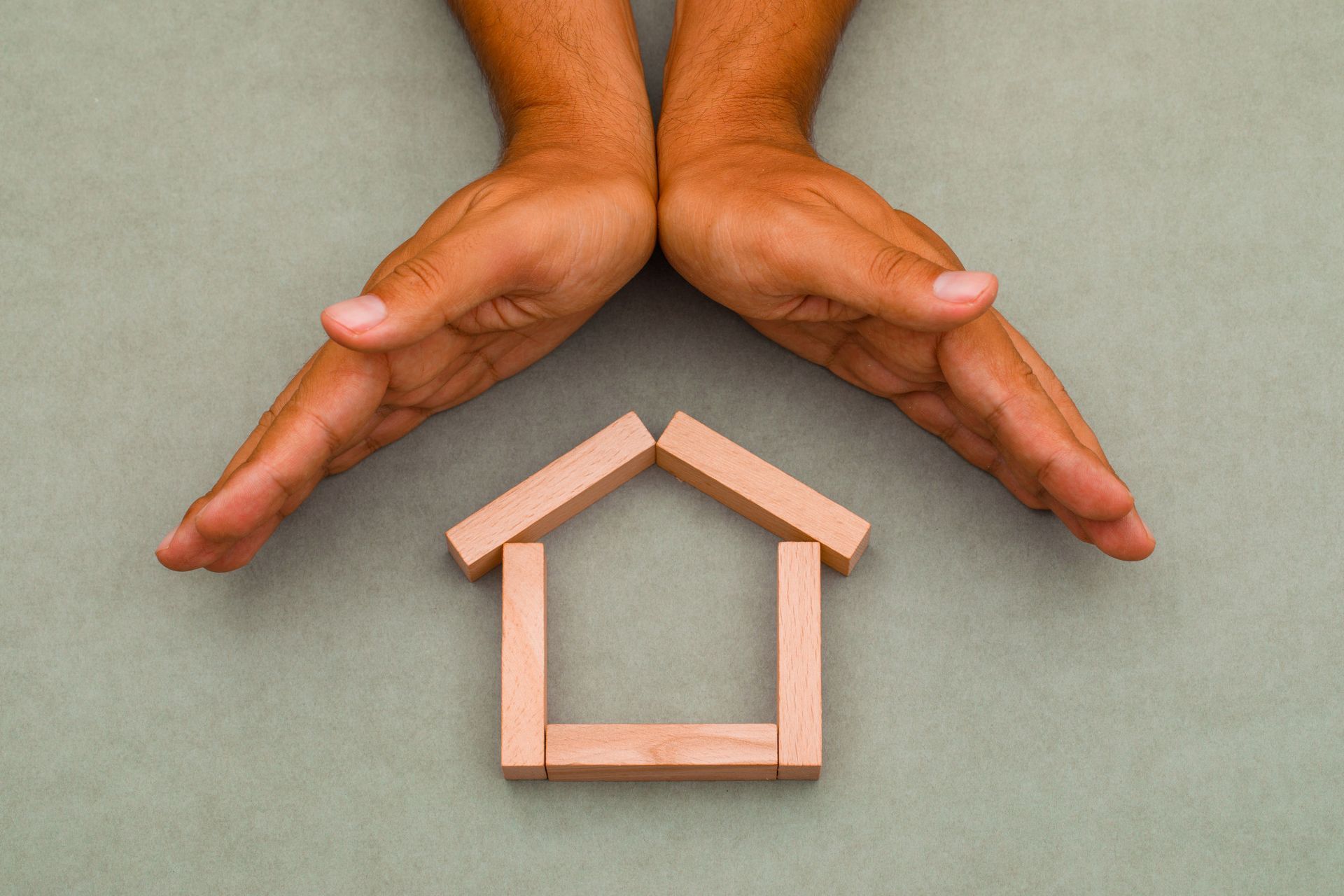 Hands forming a roof over a small wooden house shape on a gray background