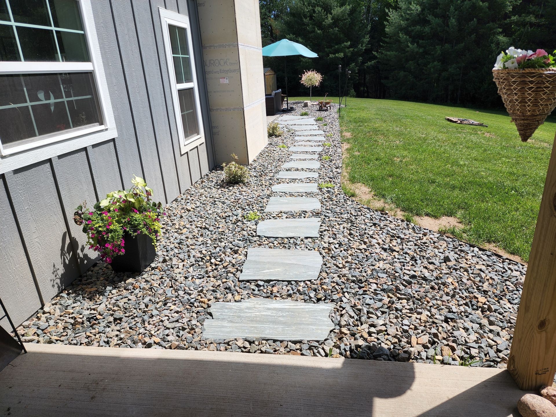 Stone path leads from porch along house to a patio with umbrella, surrounded by gravel and grass.