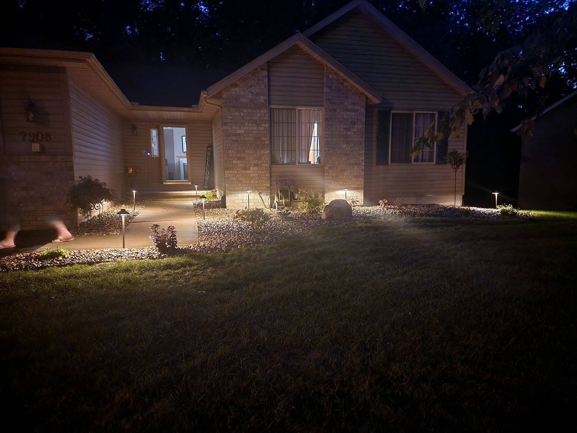 House exterior at night, illuminated by pathway lights. Brick and siding facade, glowing front door, dark sky.