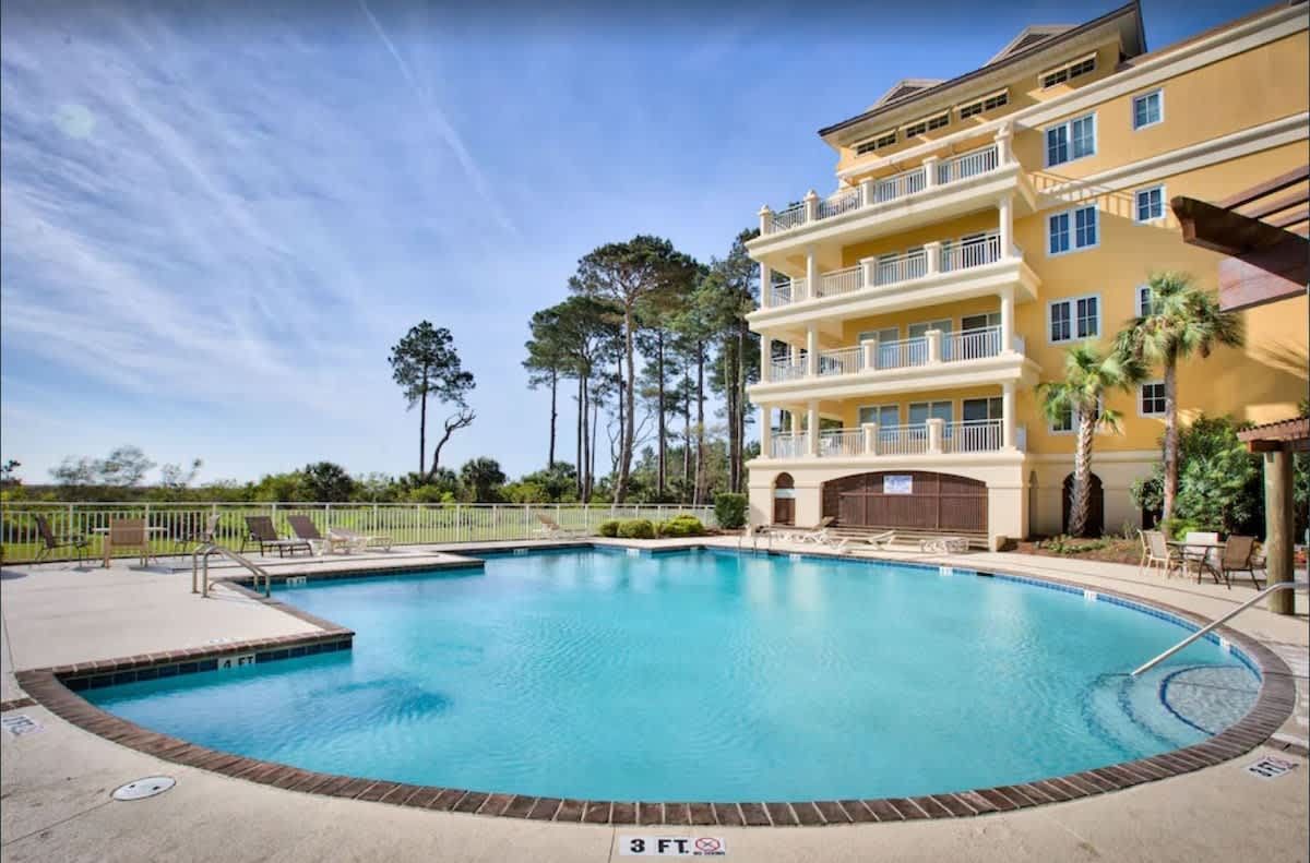 Pool and condo building on a sunny day. Blue pool, yellow building, trees in background.