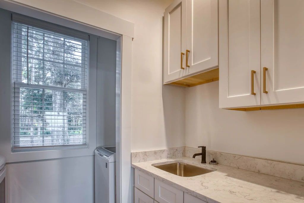 A laundry room with white cabinets , a sink , and a window.