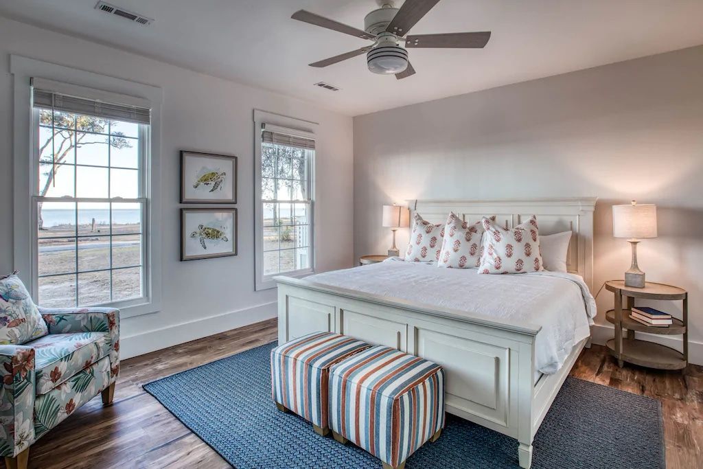 A bedroom with a king size bed , chair , ottoman and ceiling fan.