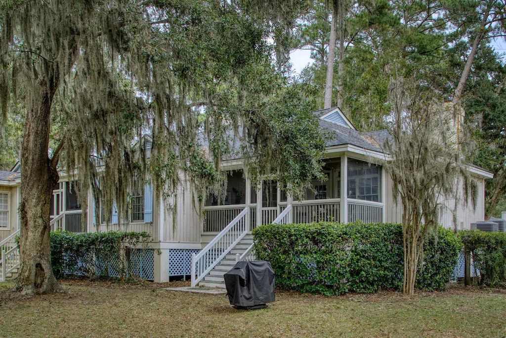 A small white house with a screened in porch is surrounded by trees and bushes.